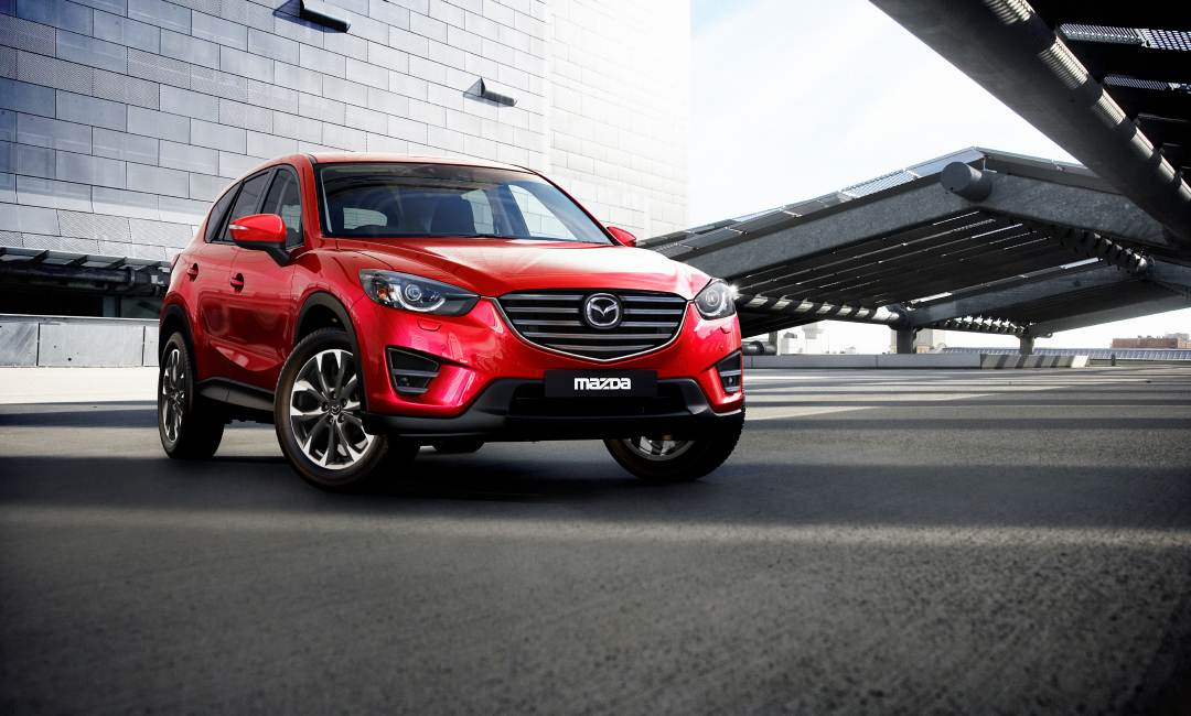 First‑generation Mazda CX‑5 in red parked in a modern urban setting, showcasing its front grille and sporty SUV design.