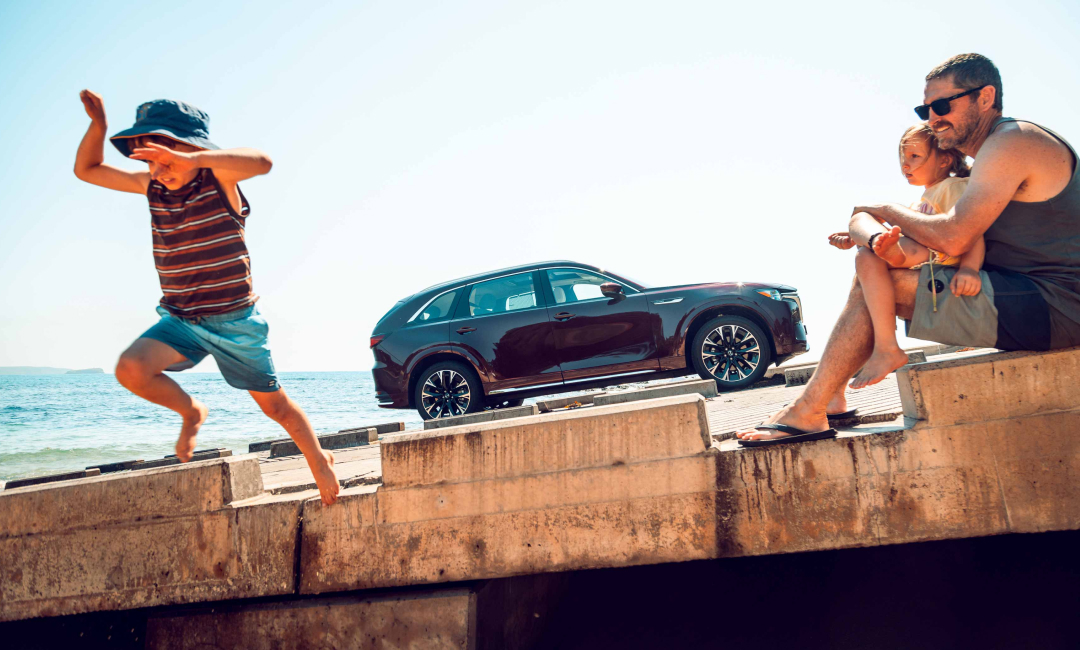 Boy leaping off a breakwater as his father and sister watch, the Mazda CX-90 parked behind