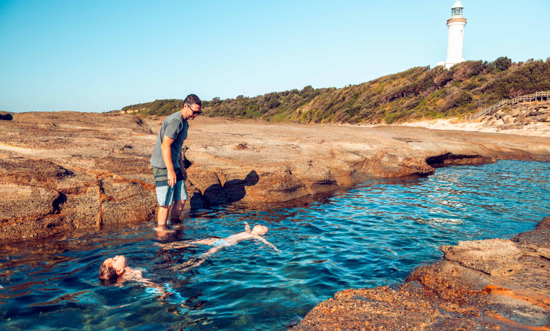 Children swimming in a coastal rockpool with Norah Head Lighthouse on the headland behind