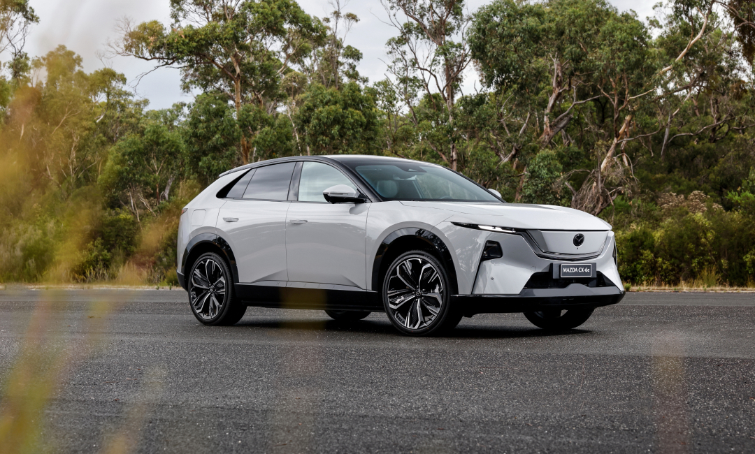 White Mazda CX‑6e electric SUV parked on a paved road with forest backdrop, showcasing modern design and alloy wheels.