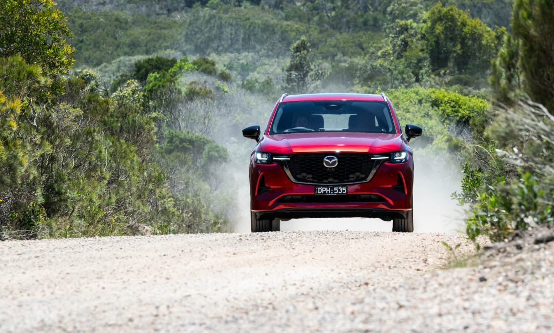 Red Mazda CX‑80 driving along a gravel road through Australian bushland, kicking up dust during a storm‑chasing journey.