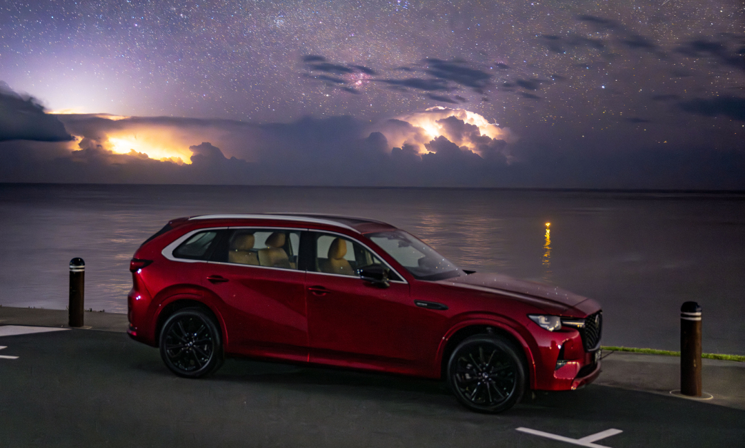 Red Mazda CX‑80 parked beside calm water at night, with lightning illuminating storm clouds beneath a star‑filled Australian sky.