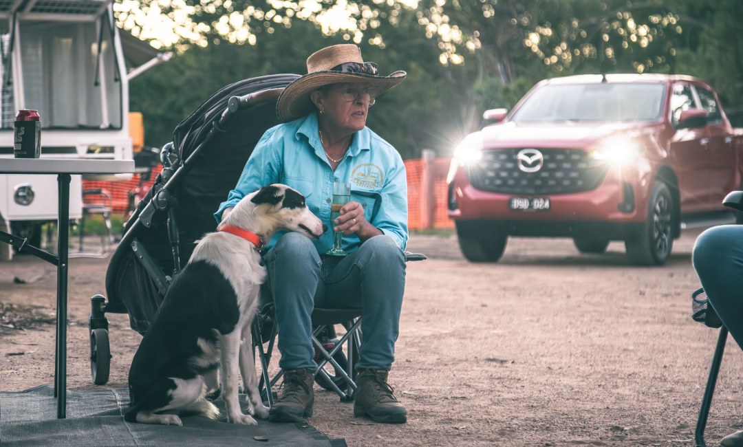 Sheepdog resting beside a seated participant at a rural campsite during Australian sheepdog trials, with a Mazda BT‑50 parked in the background.