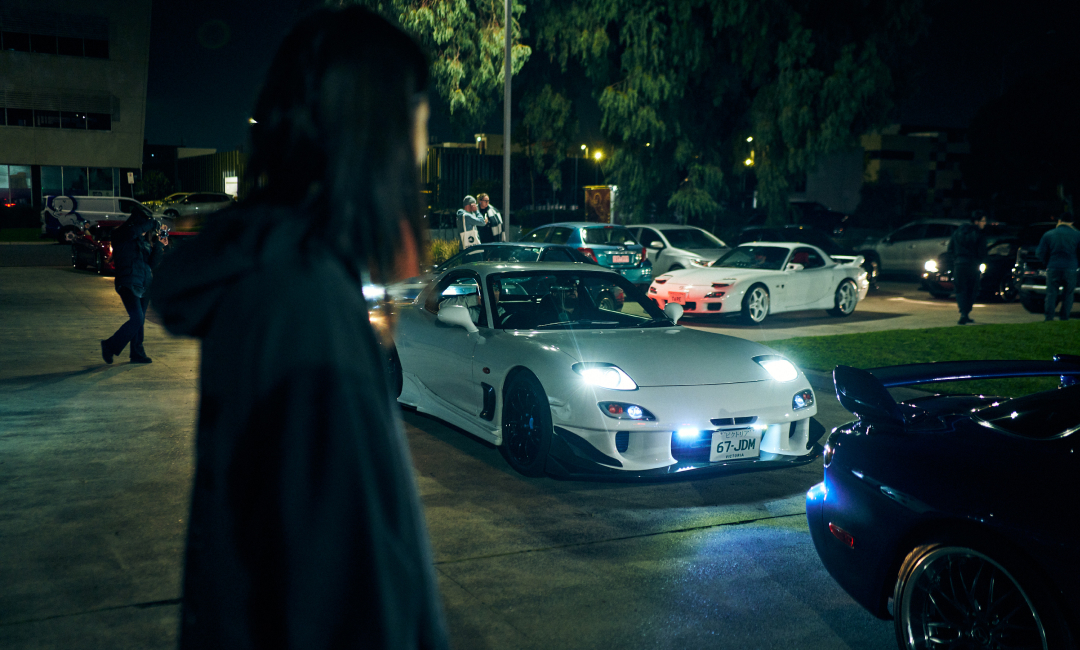 White Mazda RX-7 FD with headlights on arriving among rotary cars at night for 7's Day 2026