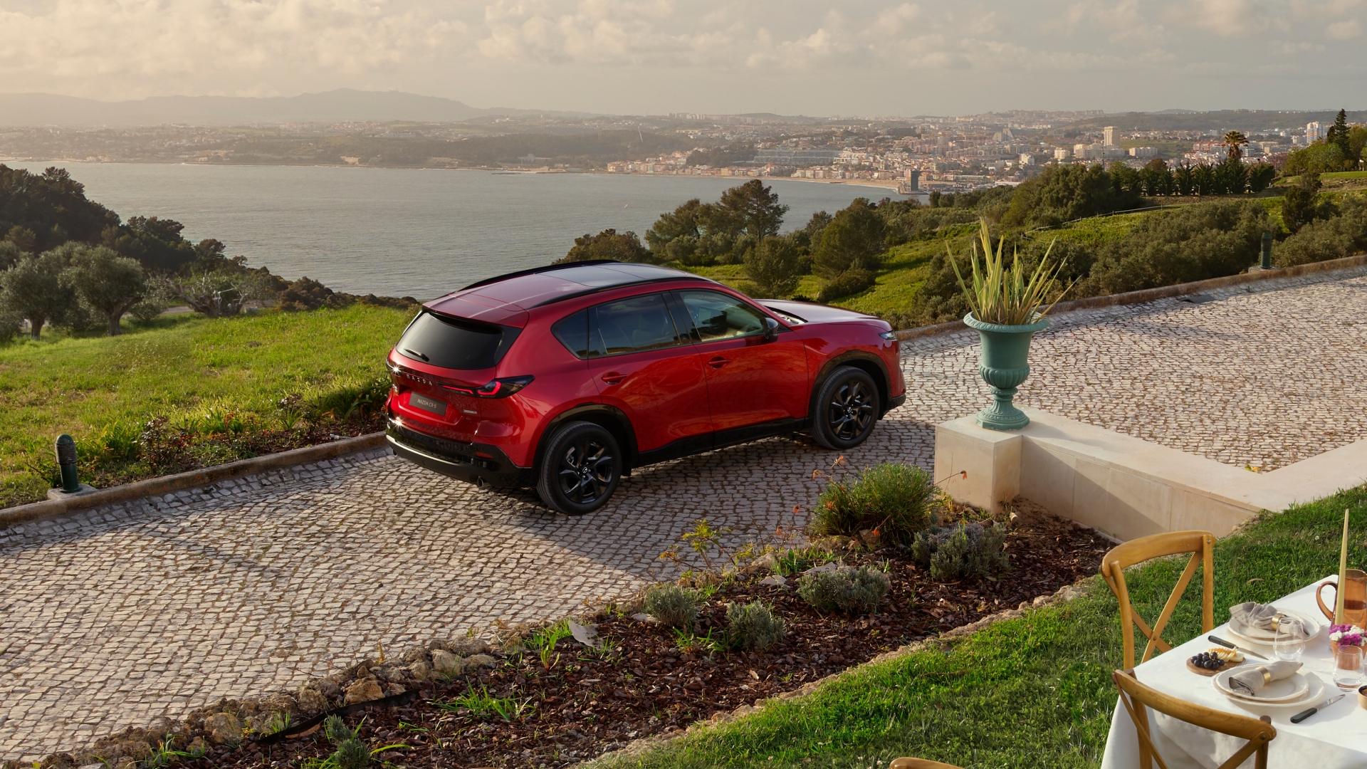 2026 Mazda CX-5 in Soul Red Crystal parked on a European cobblestone driveway with scenic sea view, black alloy wheels, and outdoor dining setup—luxury SUV lifestyle in coastal countryside setting.