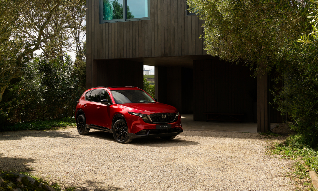 2026 Mazda CX-5 in Soul Red Crystal parked in front of a modern dark-toned house with large windows, surrounded by lush greenery and gravel driveway—stylish SUV in contemporary residential setting.