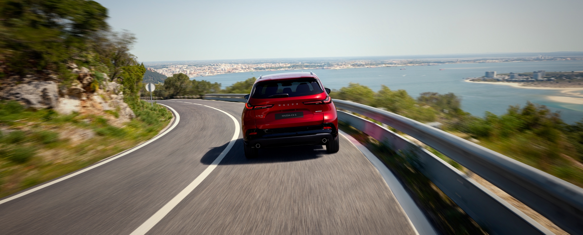 Rear of the 2026 Mazda CX-5 in Soul Red Crystal driving along a scenic coastal road with sea views, lush greenery, and distant cityscape—premium SUV performance in picturesque seaside setting.