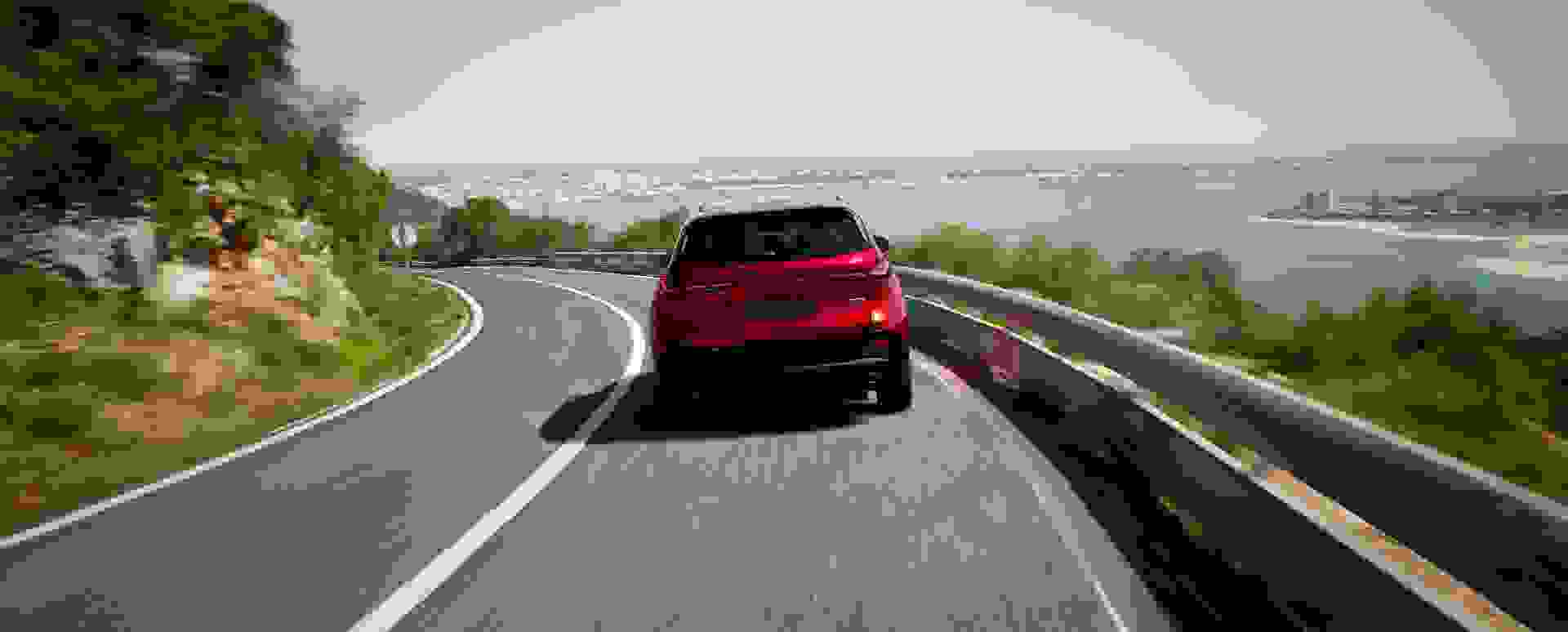 Rear of the 2026 Mazda CX-5 in Soul Red Crystal driving along a scenic coastal road with sea views, lush greenery, and distant cityscape—premium SUV performance in picturesque seaside setting.