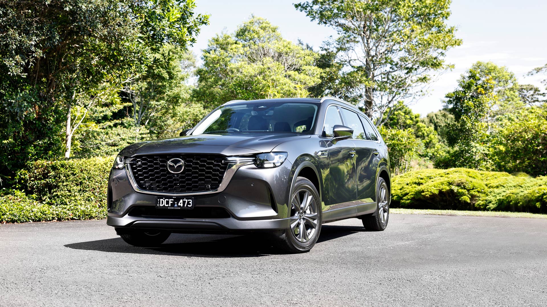 Dark grey 7-Seat Mazda CX-80 SUV parked on a paved surface surrounded by lush greenery, featuring a bold front grille, sharp LED headlights, and sleek aerodynamic design.
