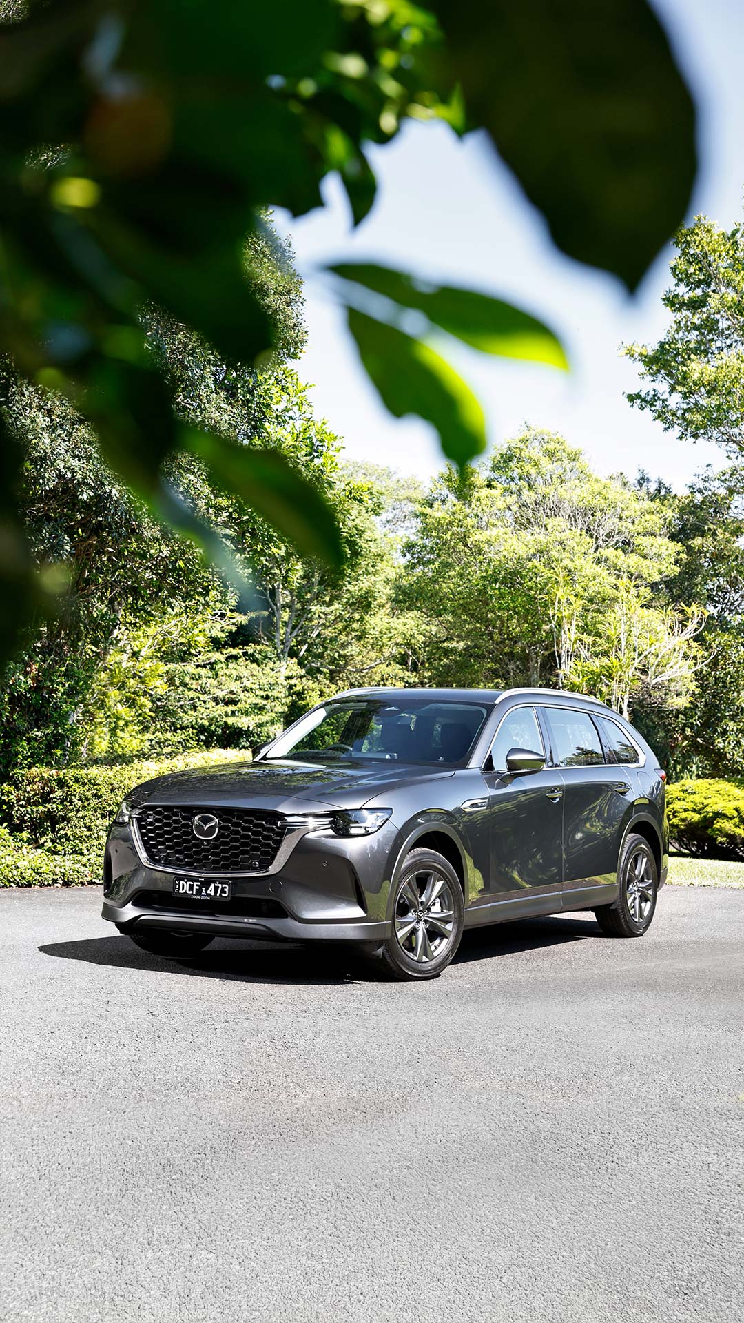 Dark grey 7-Seat Mazda CX-80 SUV parked on a paved surface surrounded by lush greenery, featuring a bold front grille, sharp LED headlights, and sleek aerodynamic design.