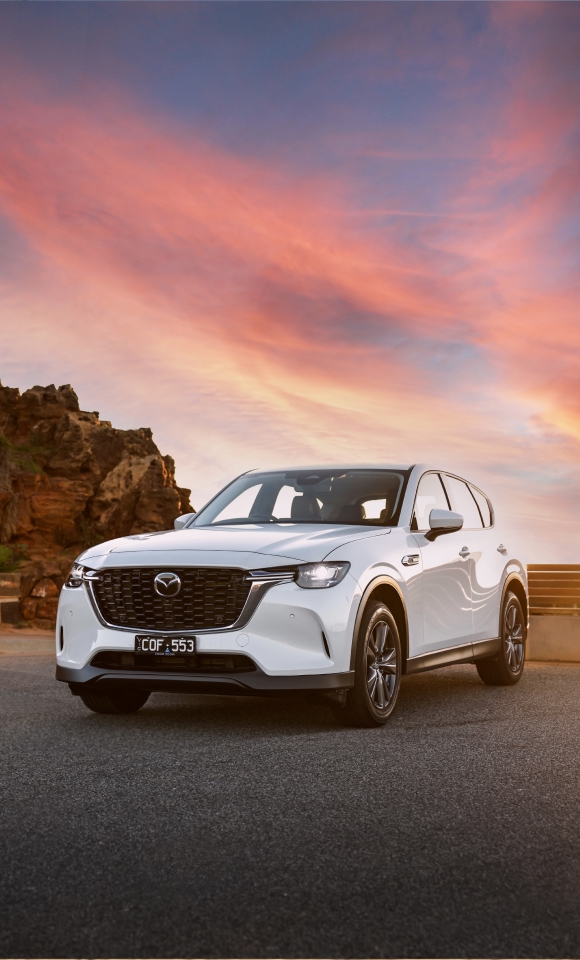 Front three-quarter view of a silver 2025 Mazda CX-60 parked on a scenic coastal road, with the ocean and cliffs in the background under a clear sky.