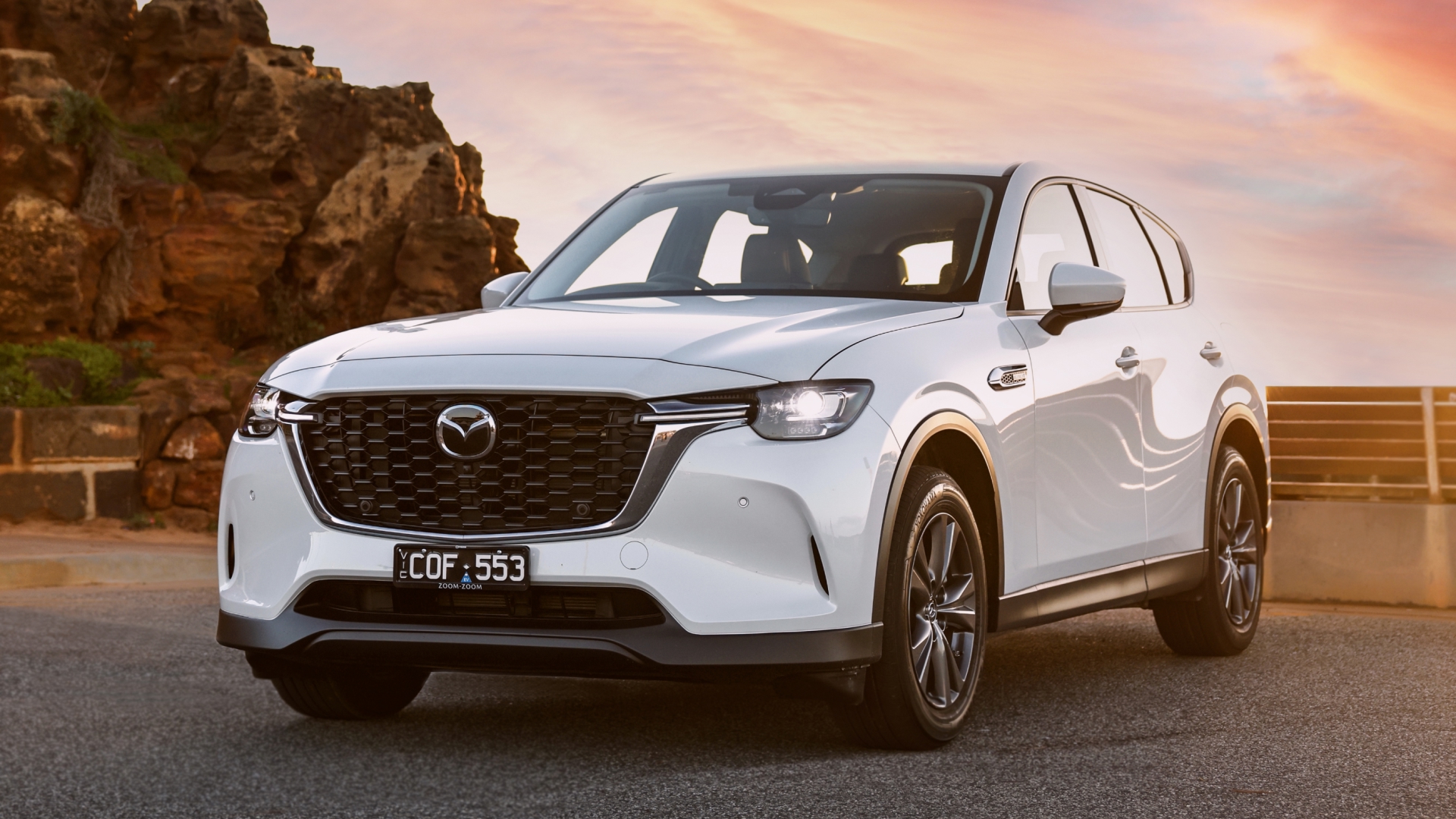 Front three-quarter view of a silver 2025 Mazda CX-60 parked on a scenic coastal road, with the ocean and cliffs in the background under a clear sky.