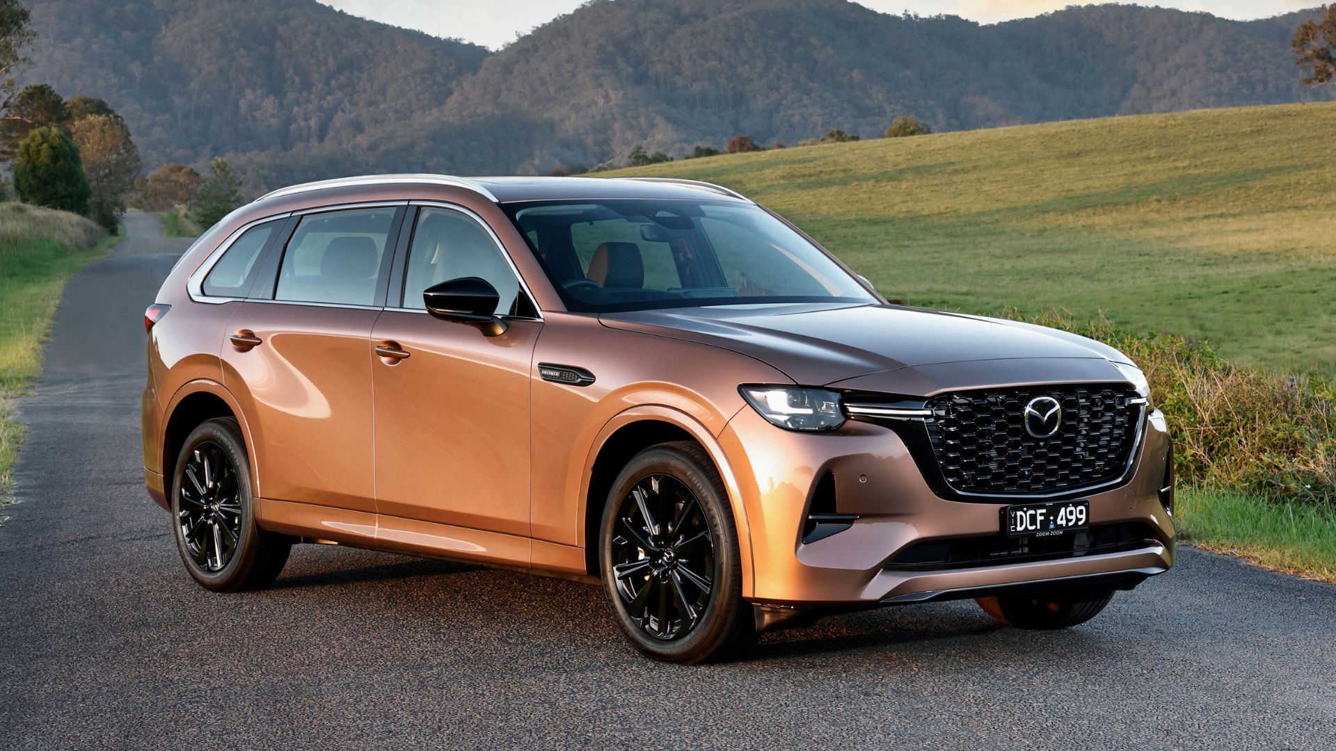Melting Copper Mazda CX-80 SUV parked on a country road with green fields and forested hills in the background, featuring sleek design, black alloy wheels, and LED headlights.