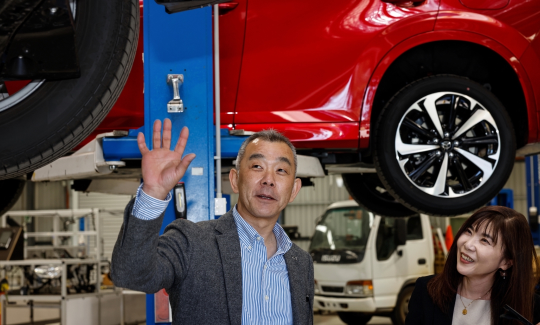A car expert in an automotive workshop demonstrates the updated suspension system of the 2025 Mazda CX-60, with a red vehicle elevated on a hydraulic lift and various tools and equipment in the background.
