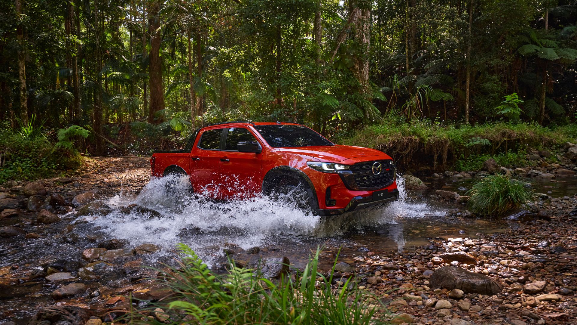 All-New 2025 Red Earth Mazda BT-50 ute driving through water, showcasing off-road capabilities in a dense tropical forest.
