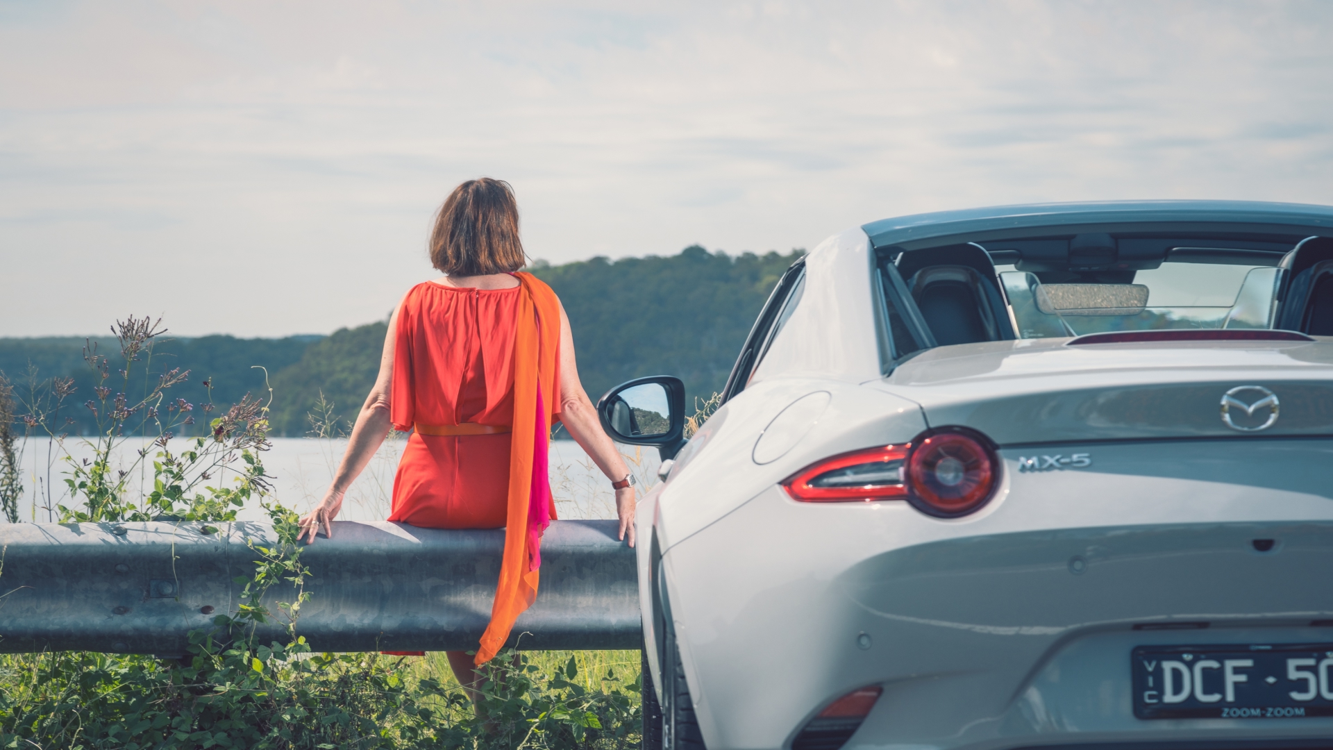 Woman sitting on a guardrail facing a lake, with a Mazda MX-5 convertible behind her.