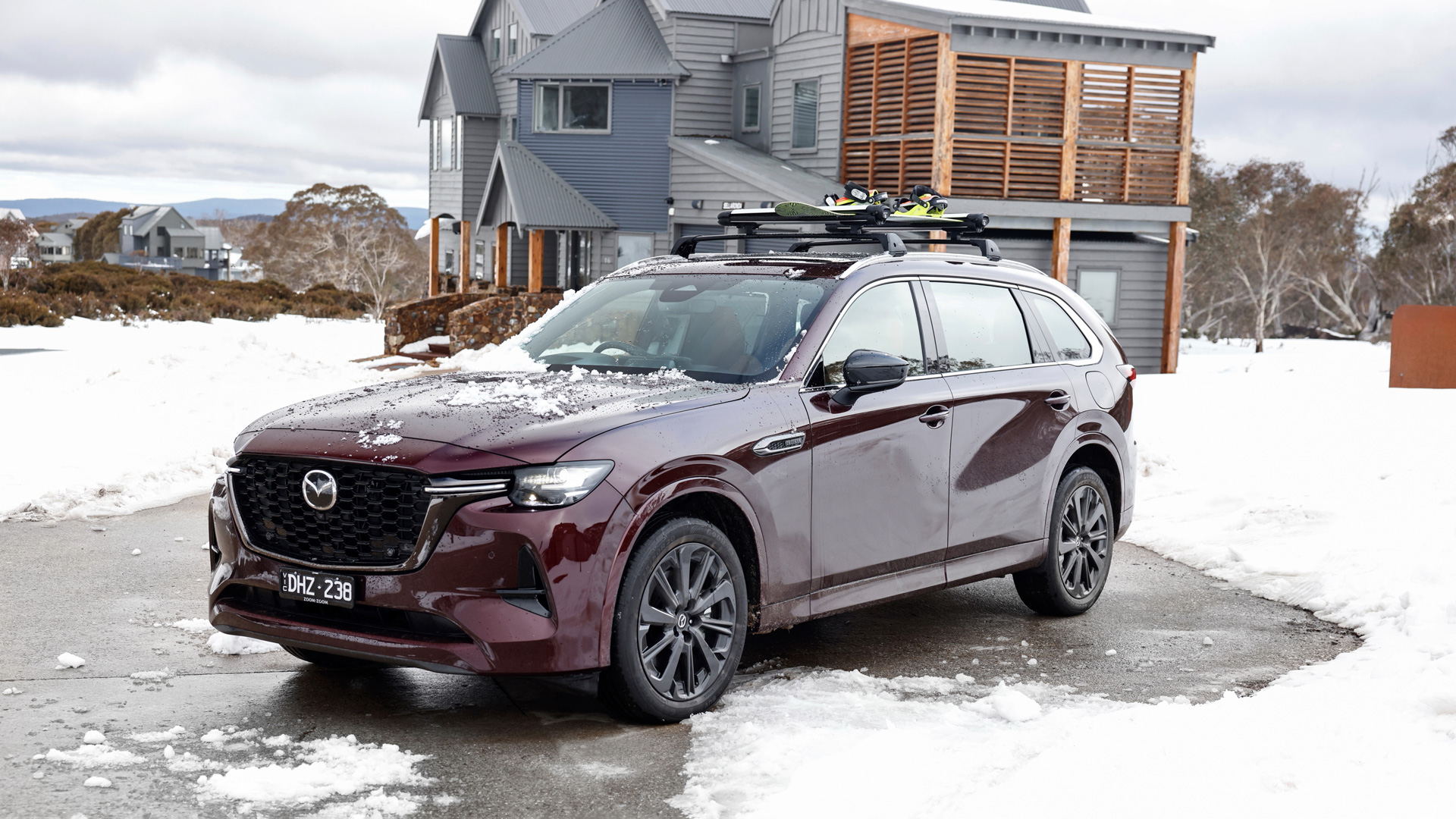 Mazda CX-80 SUV parked on a snowy driveway with ski rack, in front of modern grey chalet