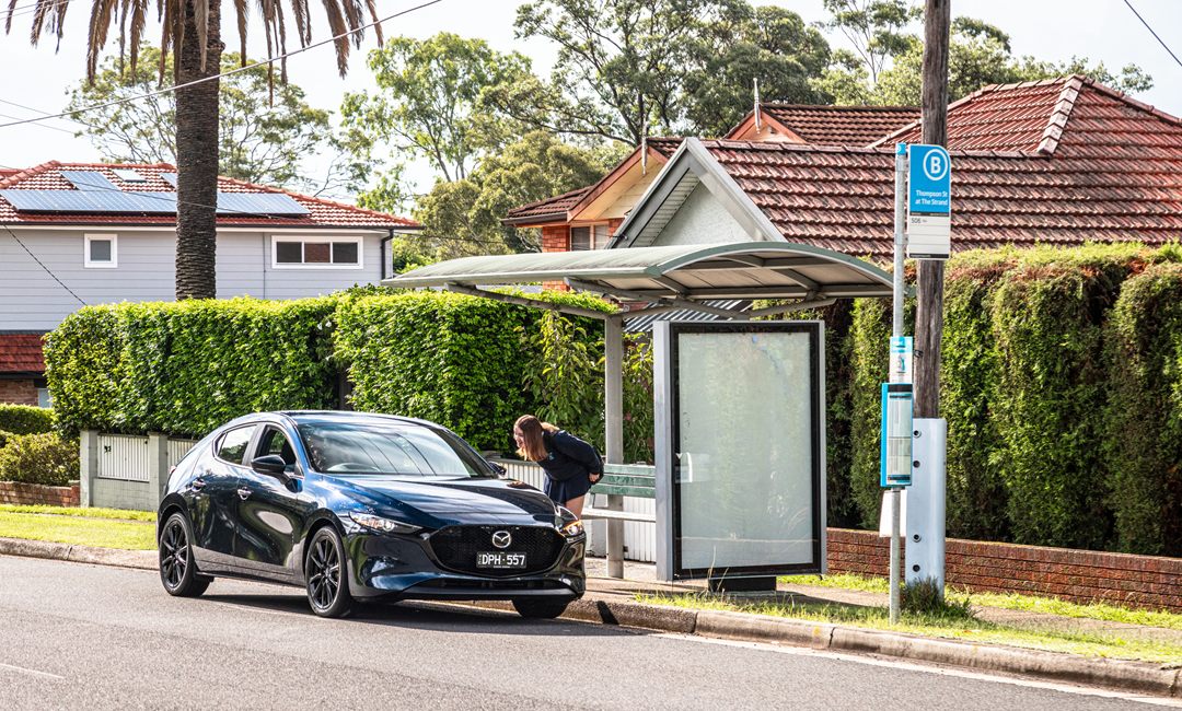 Mazda3 pulling up to collect a passenger at a suburban bus shelter