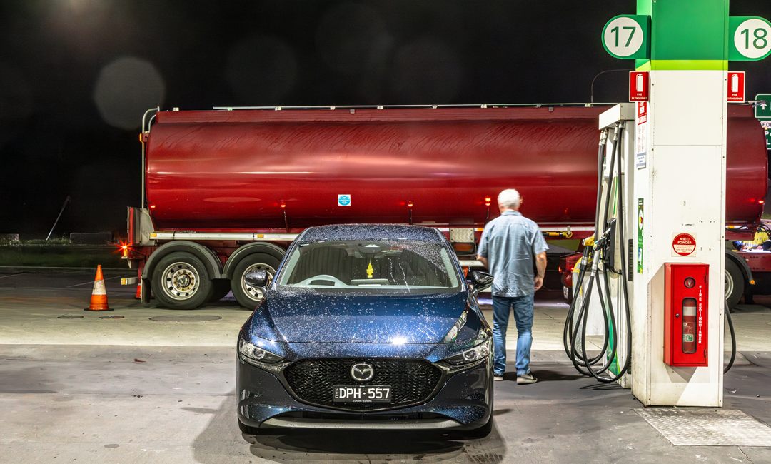 Blue Mazda3 refuelling at a service station at night beside a red fuel tanker