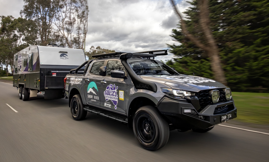 Mazda BT-50 towing a caravan along a lush, tree-lined scenic route, showcasing Snowy River Caravans and Alex's Outback Run branding &mdash; ideal for outdoor adventure and caravan lifestyle promotions.