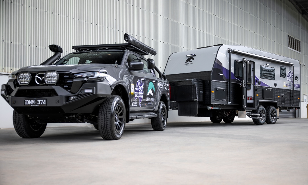 Mazda BT-50 ute towing a large camper, branded with Alex Barbas sponsor logos, parked in front of a warehouse &mdash; showcasing rugged adventure gear and caravan lifestyle branding.