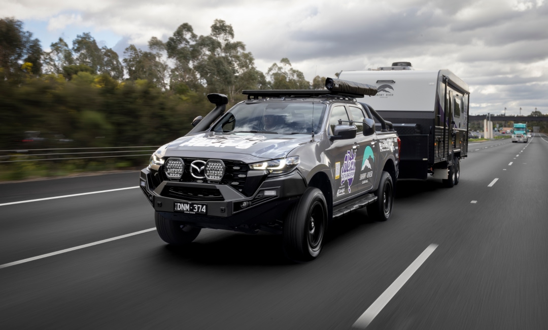 Mazda BT-50 ute towing a large camper trailer on a highway, branded with Alex Barbas sponsor logos, surrounded by trees and passing vehicles &mdash; highlighting rugged travel and caravan lifestyle