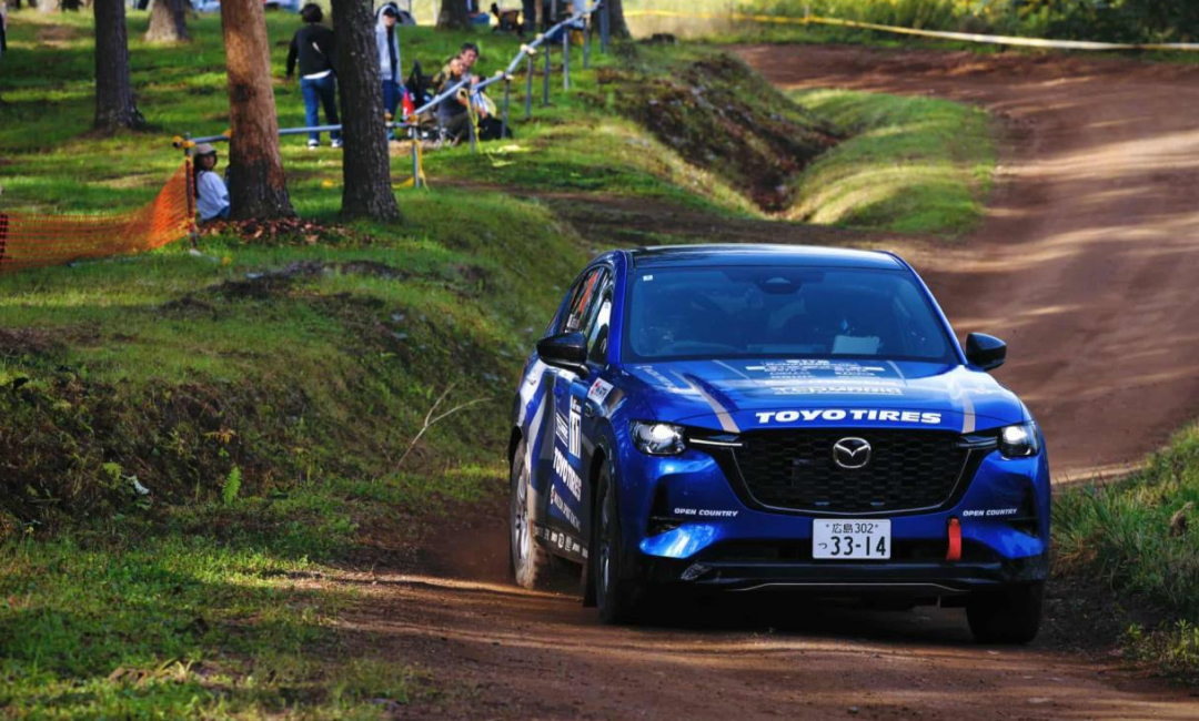 Blue Mazda CX-60 rally car powers through a dirt track at Rally Hokkaido, sponsored by Toyo Tires. Action-packed motorsport moment with spectators in the background.