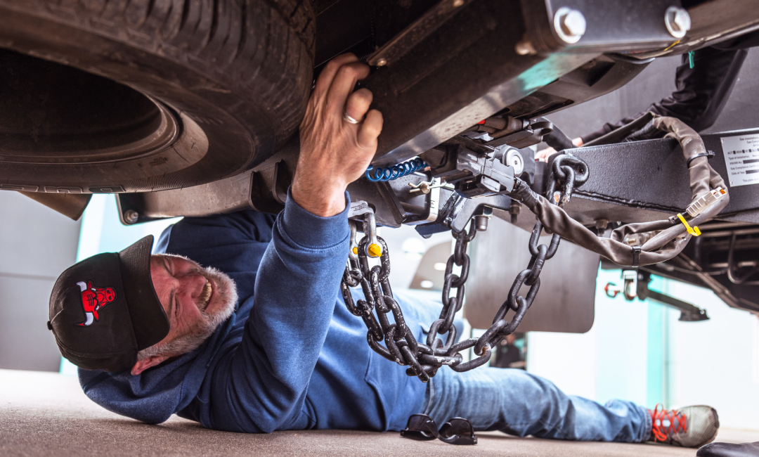 Person in blue hoodie and black cap inspecting or performing maintenance under a ute, with mechanical components and chains visible.