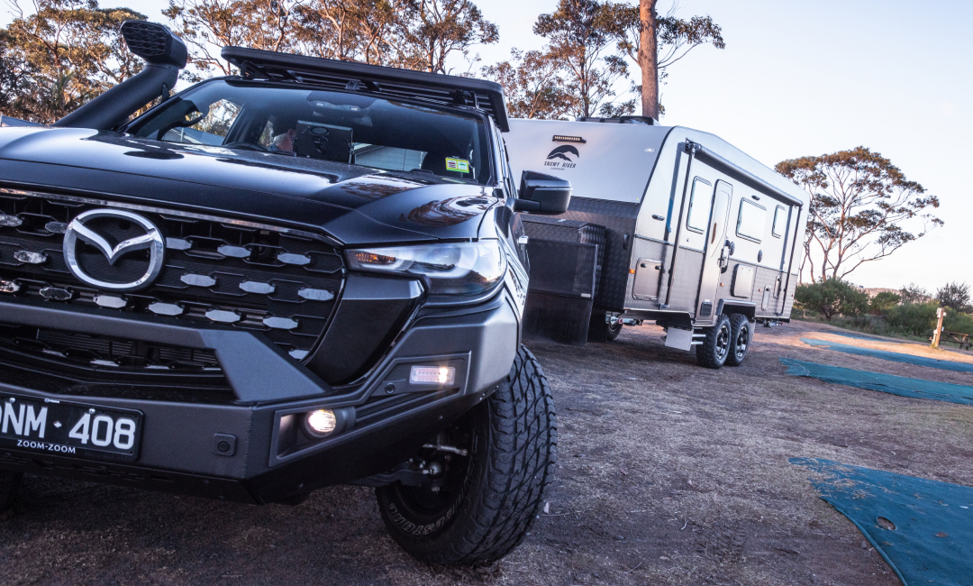 Black Mazda BT-50 pickup truck towing a large silver caravan in an outdoor setting with trees in the background, showcasing towing capability for camping or travel.