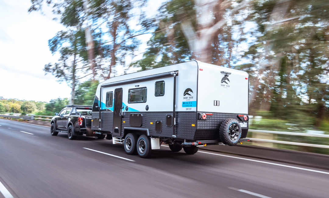 Black Mazda BT-50 towing a caravan with blue accents on a highway, surrounded by motion-blurred trees.