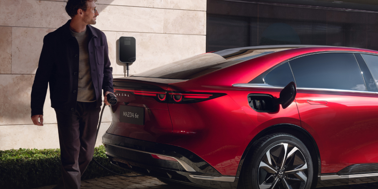 Person charging a red Mazda 6e electric car parked in front of a modern building with beige stone walls and greenery, using a wall-mounted EV charger.