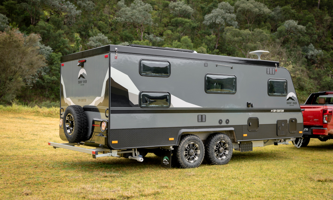 Off-road caravan with dual axles parked on grassy field near forest, attached to a red BT-50 ute.