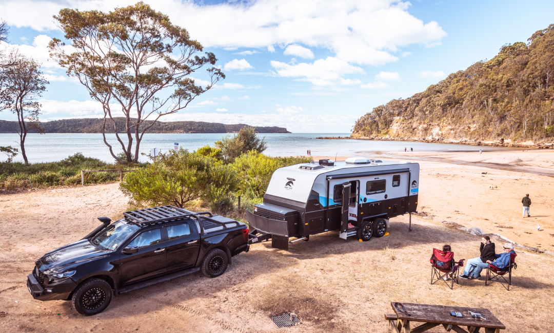 Black Mazda BT-50 towing a modern caravan on a sandy beachside campsite along the Australian coastline, with ocean views, coastal cliffs, and tall gum trees in the background; two people seated in camping chairs enjoying a summer road trip near the water.