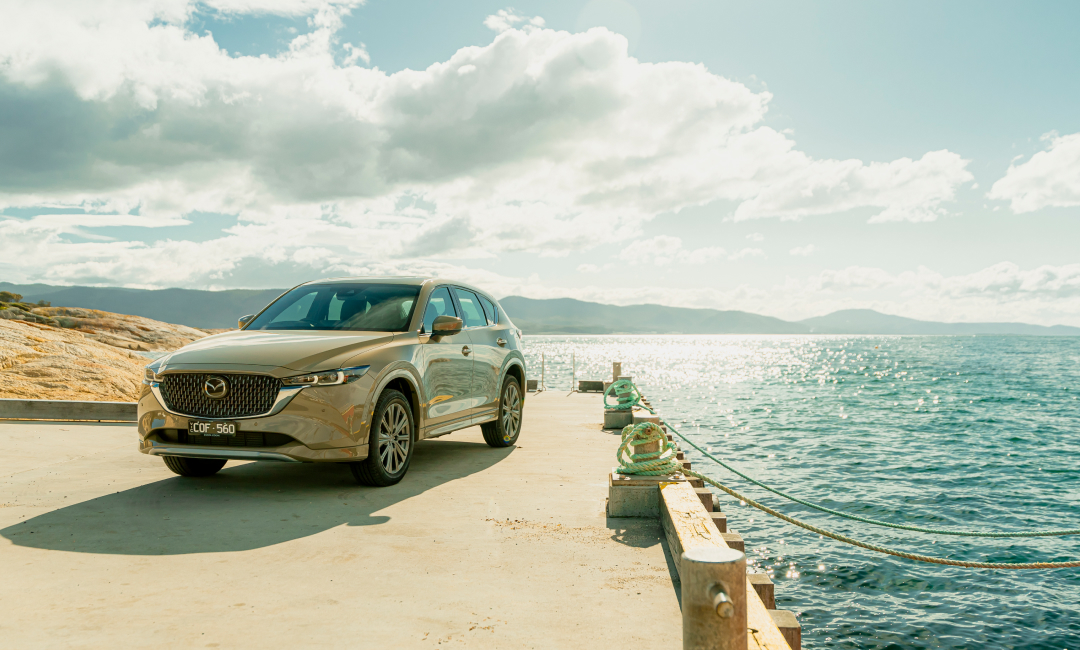 Mazda CX-5 parked on a concrete pier overlooking sparkling blue ocean waters under a bright sunny sky, with rugged coastal cliffs and distant mountains in the background, showcasing an Australian seaside road trip destination.