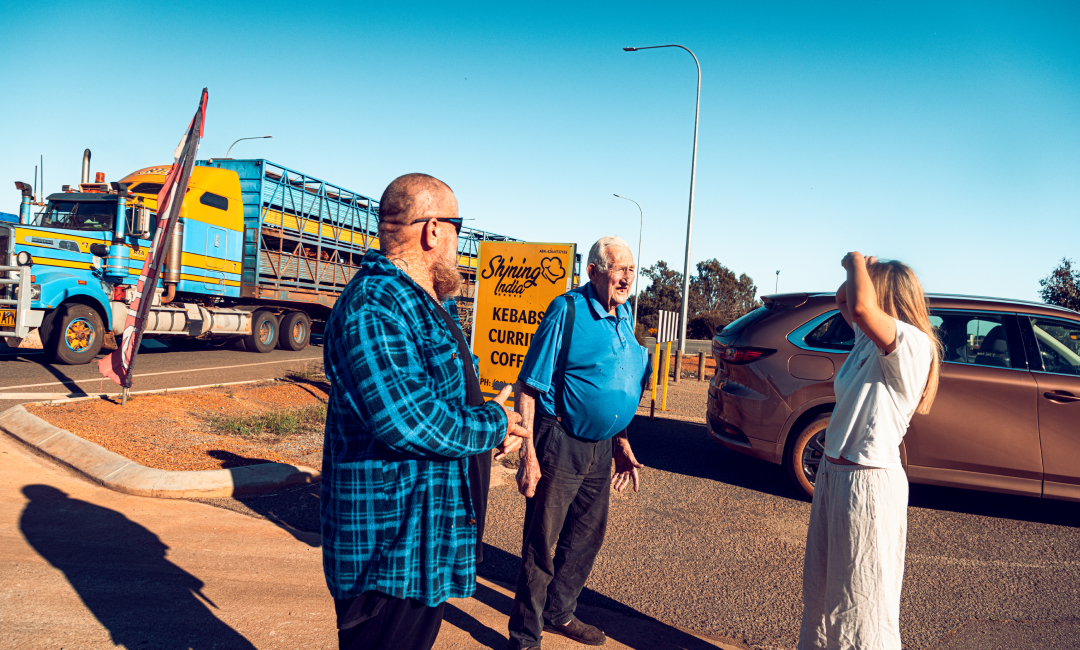 Mazda CX-80 in Melting Copper parked at a roadside stop in Western Australia during a wildflower road trip, traveler engaging with locals and discussing wildflower locations, with a colorful livestock truck passing in the background under clear blue skies.